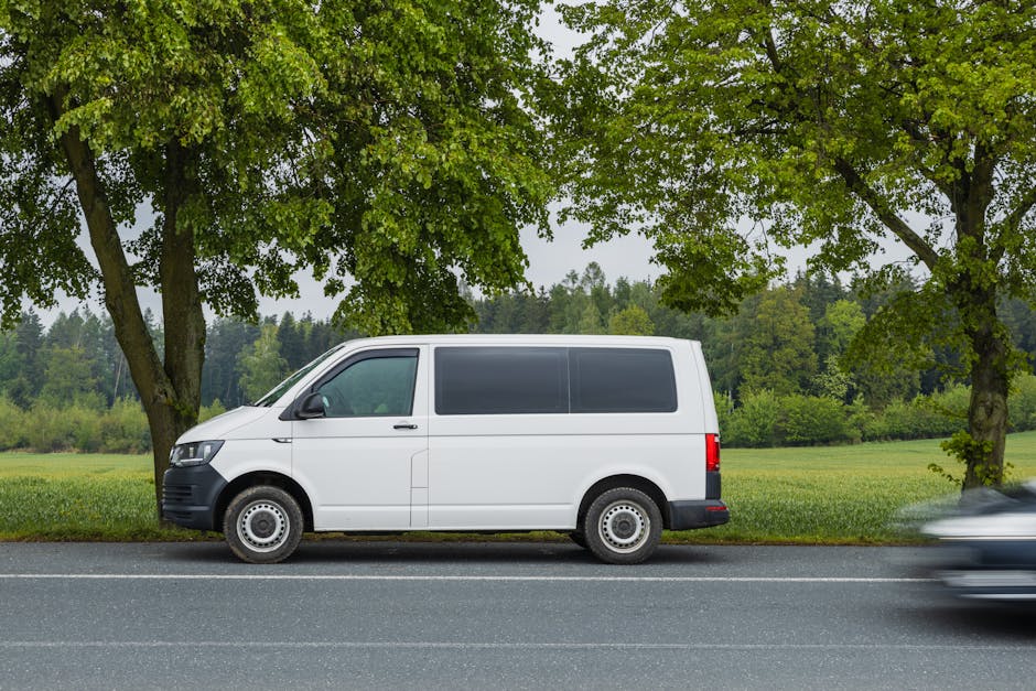 A white panel van parked parallel along a quiet road in Ardleigh Green, with two tall deciduous trees with green foliage and a grassy field in the background. The van is part of a home relocation or furniture transport process managed by Man with Van Ardleigh Green, a removal service specialist. The vehicle has tinted rear windows, black wheel covers, and a black bumper. The setting appears to be during daylight hours, under natural lighting, with the van positioned close to the curb, ready for loading or unloading furniture, boxes, and packing materials as part of a house move. A slight motion blur is visible on a vehicle passing in the foreground, indicating movement along the street. This scene captures the logistics involved in packing, loading, and transporting belongings to support a house relocation or removal service.