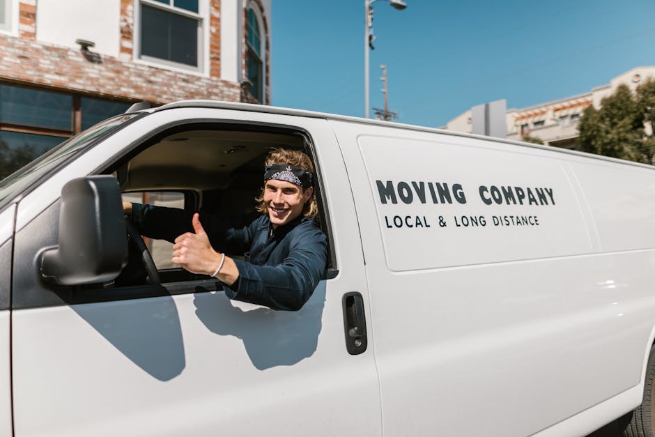 A smiling young man with long hair, wearing a black headband and dark clothing, is sitting in the driver's seat of a white moving van labeled 'MOVING COMPANY, LOCAL & LONG DISTANCE,' inside a property during daylight. The man is giving a thumbs-up with his right hand through the open window while looking towards the camera. The van is parked on a paved surface, nearby a brick building with large glass windows and residential structures, indicating a home relocation scene. In the background, a blue sky with some clouds is visible, along with utility poles and greenery. The scene captures the loading process, typical of professional removals by Man with Van Ardleigh Green, showing the vehicle ready for furniture transport and packing and moving activities during home moving logistics.