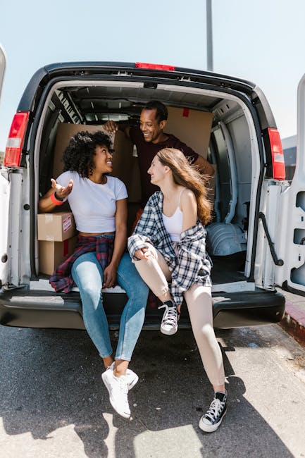 Two young women sit on the edge of an open moving van, engaged in conversation, with a man standing inside the vehicle smiling. The van's interior shows cardboard boxes, some wrapped in plastic or packing paper, and pieces of furniture covered with protective blankets. The scene takes place outdoors on a paved driveway, with sunlight illuminating the scene. The women are casually dressed, one in jeans and a t-shirt, the other in leggings and a plaid shirt tied around her waist. The man is dressed in a dark shirt, assisting with home relocation preparations. Visible equipment includes a trolley or dolly near the entrance of the van, and the surrounding environment suggests a residential area. This image depicts the loading process during a house move, with furniture and packing materials being organized for transport, aligning with professional removals and moving services provided by Man with Van Ardleigh Green.