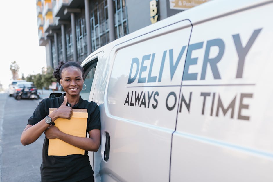 A smiling young man with long hair, wearing a black headband and dark clothing, is sitting in the driver's seat of a white moving van labeled 'MOVING COMPANY, LOCAL & LONG DISTANCE,' inside a property during daylight. The man is giving a thumbs-up with his right hand through the open window while looking towards the camera. The van is parked on a paved surface, nearby a brick building with large glass windows and residential structures, indicating a home relocation scene. In the background, a blue sky with some clouds is visible, along with utility poles and greenery. The scene captures the loading process, typical of professional removals by Man with Van Ardleigh Green, showing the vehicle ready for furniture transport and packing and moving activities during home moving logistics.