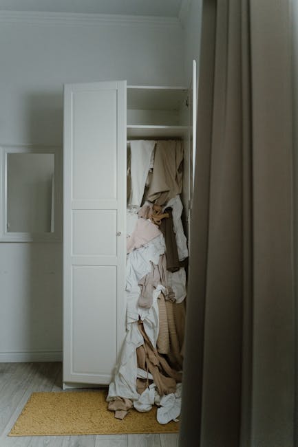 An open white wardrobe with multiple shelves and door panels, positioned against a light-colored wall inside a room, with a large pile of crumpled and folded clothing in various shades of beige, white, and brown spilling out and occupying part of the floor in front of it. The wardrobe appears to be part of a house relocation or packing process, with the clothing possibly prepared for removal or moving. To the right, partially visible, is a grey curtain hanging beside the wardrobe, and the floor is covered with a light wooden or laminate surface with a small yellowish rug underneath the wardrobe. The scene is lit with natural light, and the setting suggests an interior space being prepared for furniture transport or home relocation, supported by services such as those from Man with Van Ardleigh Green.