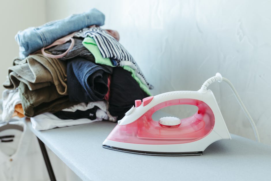 A pile of folded clothes in various colours and fabrics, including cotton and striped patterns, placed on a white ironing board. Next to the clothes, there is a modern steam iron with a white and pink design, plugged in and ready for ironing. The scene appears to be set inside a home, indicating laundry or packing activities associated with home relocation. The ironing board is situated against a light-colored wall with natural light illuminating the area. This setup reflects typical household chores involved in preparing items for a move, aligning with house removals and packing services offered by Man with Van Ardleigh Green. The arrangement of items suggests an environment focused on packing, organizing, and furniture transport as part of a comprehensive moving process involved in furniture and appliance handling during a house move.