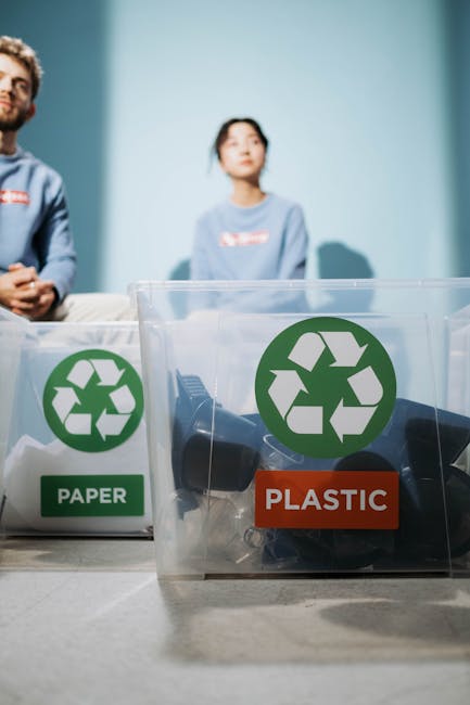 Close-up of two transparent recycling bins placed on a white surface in an indoor setting, each with a green recycling symbol. The left bin is labelled 'PAPER' and contains various paper waste, while the right bin is labelled 'PLASTIC' and holds black plastic items and packaging materials. In the background, two people, a man and a woman, are seated against a light blue wall, slightly out of focus, with their upper bodies visible. The scene emphasizes environmentally conscious waste separation, relevant to eco-friendly removals and recycling practices in house moving situations, with the bins positioned in the foreground and the individuals involved in packing or sorting tasks behind.