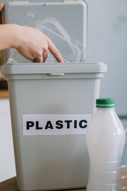 A close-up view of a grey plastic recycling bin with a white label that reads 'PLASTIC' in bold black letters, situated on a flat surface indoors. The bin's lid is open, revealing clear plastic bottles inside, with one finger visible pointing towards them. To the right of the bin, there is a partially visible white plastic bottle with a green cap. The setting appears to be in a home environment, possibly during a packing or disposal process as part of home relocation or house removals. This image supports the concept of eco-friendly recycling within the context of a house move, consistent with services provided by Man with Van Ardleigh Green.