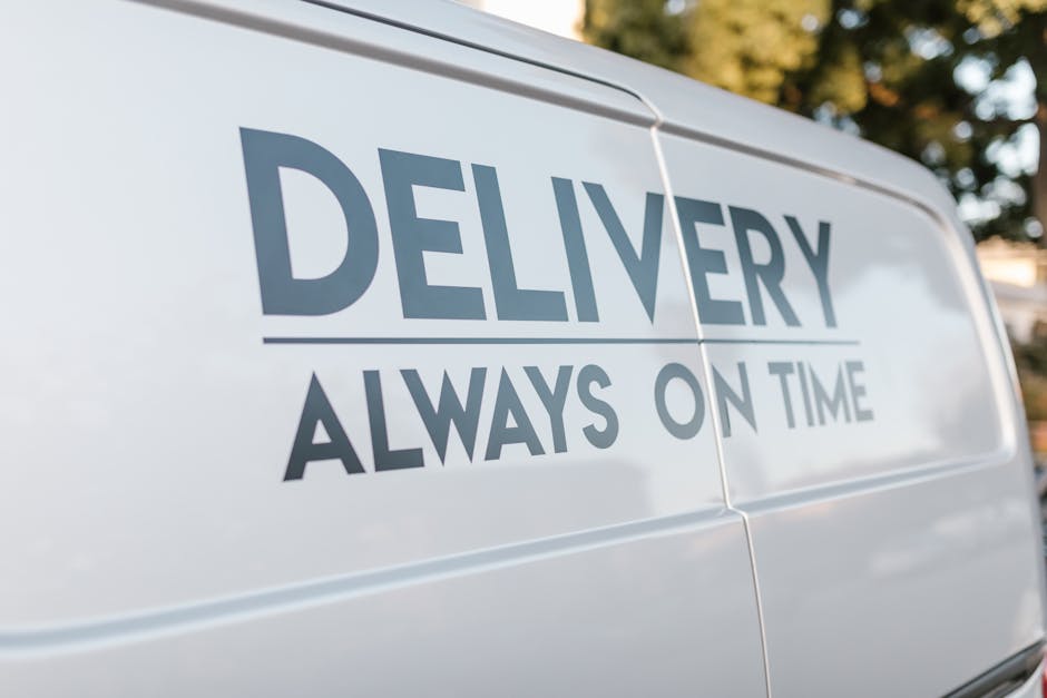 Close-up image of a white delivery van parked outdoors, with the company slogan 'Delivery Always on Time' prominently displayed in large, dark blue letters on the side panel. The van appears to be part of a professional removals service, with a focus on reliable furniture transport and home relocation. The background features blurred green foliage and hints of sky, indicating daylight conditions. The van's surface is clean and reflective, emphasizing a well-maintained vehicle used for packing and moving logistics. Inside the image, no personnel or objects are visible, solely the branding and exterior of the van, which aligns with the theme of professional house removals and transport services provided by Man with Van Ardleigh Green, especially relevant to navigating house removal permits or parking regulations for Ardleigh Green relocations.