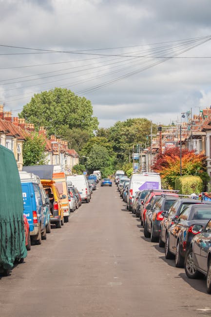 A residential street scene during daytime showing a narrow, paved road lined with closely parked cars on both sides, including hatchbacks, sedans, and vans. Several cardboard boxes, wrapped furniture pieces with protective blankets, and plastic-wrapped household items are visible on the pavement, indicating a home relocation process. A man with a van from Man with Van Ardleigh Green is partially visible, possibly in the process of loading or unloading furniture and packing materials. The background features terraced houses with brick facades, tiled roofs, and small front gardens with trees and bushes, set against an overcast sky with grey clouds. Power lines run overhead, and some houses display house removal permits or parking notices, consistent with controlled parking zones related to shifting home contents within the area. The image captures the logistical elements of furniture transport and packing during an internal move, emphasizing careful handling and organized load preparation within an urban neighbourhood context.