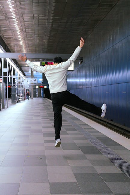 A man dressed in a white shirt and black trousers is captured mid-air inside a modern train station, with arms and legs spread wide in a dynamic pose. The station features a high, dark ceiling with a metallic finish and a wide tiled floor. Behind the man, a dark blue wall extends along the platform, with a small section of train tracks visible at ground level. There are a few people in the background, some standing near the station entrances, and digital displays hanging from the ceiling show departure information. The scene appears to depict a moment of lightheartedness or celebration during a home relocation or moving process, with the man possibly preparing to load or unload boxes or furniture as part of packing and moving activities. The environment is well-lit with a clean, orderly appearance typical of urban transport hubs, and the details suggest an indoor setting related to furniture transport or house removals undertaken by [COMPANY_NAME].
