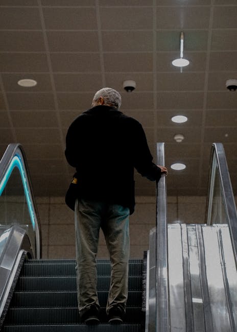 A man with grey hair wearing a black jacket and grey trousers stands on an escalator inside a station, holding onto the side rail with his right hand while looking upwards. The escalator has black steps with metal edges, and the surrounding environment features a ceiling with evenly spaced circular lights. There are visible security cameras and smoke detectors mounted on the ceiling. The setting suggests a public transport or station environment, which could relate to the process of home relocation or moving logistics, as seen in services provided by Man with Van Ardleigh Green. The image depicts an indoor scene with neutral lighting and a focus on the man's movement through the station, aligning with themes of packing and moving processes involved in furniture transport and house removals.
