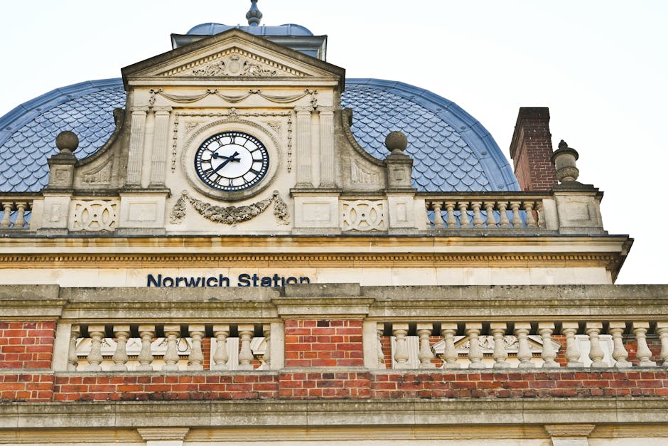A close-up view of the ornate facade of Norwich Station, featuring a central clock with black Roman numerals and clock hands set against a white background, mounted above the station’s name signage.