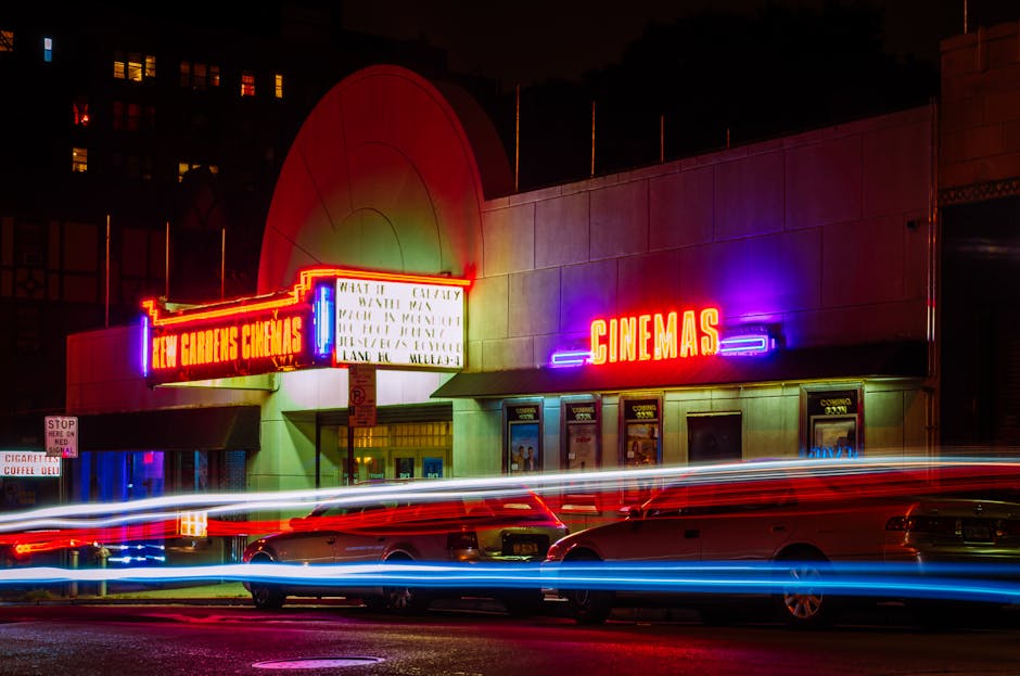 Nighttime exterior view of Kew Gardens Cinemas, with illuminated neon signs displaying 'Kew Gardens Cinemas' and 'CINEMAS' in bright red and purple colors. The building has a modern, stucco façade with large entrance doors and posters visible in the ticket window area. Multiple cars are parked in front on the street, and light trails from passing vehicles are captured due to long exposure, creating streaks of red, white, and blue across the image. The scene is lit by the cinema's neon signage and ambient street lighting, with a dark sky overhead. This setting illustrates an urban environment suitable for house removals and local relocations, captured in a way that emphasizes moving logistics and transportation considerations, relevant to services like those provided by Man with Van Ardleigh Green.