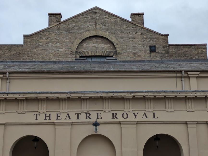 Photograph of the exterior of a historic building with a beige façade and classical architectural features, displaying the sign 'THEATRE ROYAL' above the entrance. The upper section shows a brick gable wall with a semi-circular window and a small ventilation opening. The entrance area features three arches, with the central arch fitted with a black lantern-style light fixture. The scene is set during daylight with an overcast sky, and the visible part of the building suggests it may be a location for local theatre performances. This image relates to house removals or relocation services, as provided by Man with Van Ardleigh Green, focusing on moving or accessing historic or public buildings in the context of local relocations near Queen's Theatre Hornchurch, including packing, loading, and furniture transport processes.