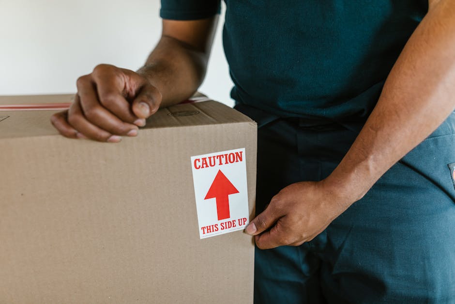 A person dressed in blue clothing is seen holding a large cardboard box during a home relocation process, with a red and white caution label indicating 'THIS SIDE UP' and an upward arrow. The individual’s hands are gripping the box, which is positioned on a flat surface indoors, likely on a trolley or dolly. The background suggests an interior setting, with part of a plain wall visible. The scene captures the careful handling and packaging aspects involved in furniture transport and packing for a local moving service, consistent with the offerings of Man with Van Ardleigh Green in their removals category. The lighting appears natural and even, emphasizing the importance of safety and proper guidance when loading and moving household items.