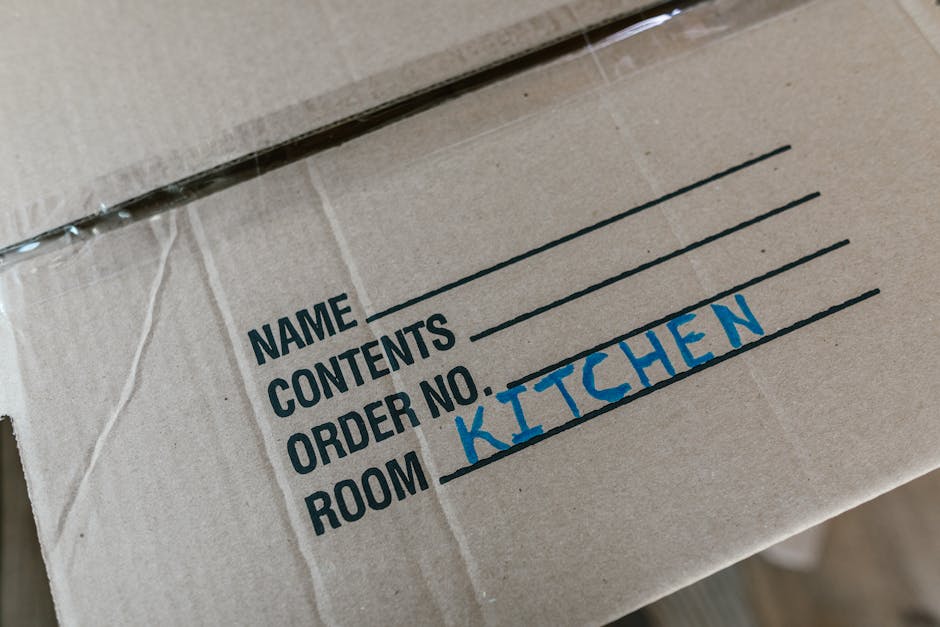 A person dressed in blue clothing is seen holding a large cardboard box during a home relocation process, with a red and white caution label indicating 'THIS SIDE UP' and an upward arrow. The individual’s hands are gripping the box, which is positioned on a flat surface indoors, likely on a trolley or dolly. The background suggests an interior setting, with part of a plain wall visible. The scene captures the careful handling and packaging aspects involved in furniture transport and packing for a local moving service, consistent with the offerings of Man with Van Ardleigh Green in their removals category. The lighting appears natural and even, emphasizing the importance of safety and proper guidance when loading and moving household items.