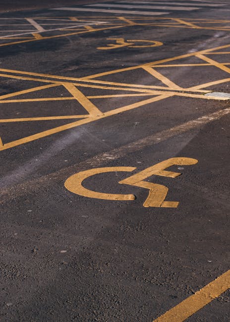 A close-up view of a commercial parking lot featuring multiple designated parking spaces marked with yellow lines and symbols. Prominently visible are two spaces designated for disabled parking, with the universal wheelchair symbol painted in yellow on the dark asphalt surface. The parking area appears to be outdoors, with the surface showing signs of wear and slight discoloration. The clear yellow markings provide guidance for vehicle parking and accessibility compliance, supporting the logistics involved in home relocation or furniture transport services. The image emphasizes efficient parking management relevant to house removals, such as the process of loading and unloading moving vehicles, and the importance of dedicated accessible parking spots for ease of moving household items. This setting aligns with the logistical considerations addressed by Man with Van Ardleigh Green when planning for parking permits and access during property removals.