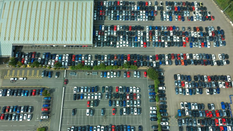An aerial view of a large parking lot adjacent to a commercial building with a flat, beige roof. The parking area contains numerous vehicles, predominantly small cars in various colours including white, red, blue, and black, parked in orderly rows. There are designated parking bays marked with white lines and a few vacant spaces visible. Near the building's entrance, a small section of yellow-striped loading zone is present, with a white van parked nearby. The parking lot is bordered by greenery and a sidewalk, with some trees and bushes along the perimeter. The scene captures the typical environment for a busy retail or office area, relevant to home relocation services such as loading, unloading, and transporting household items. Man with Van Ardleigh Green occasionally offers clearance for parking permits and managing delays during house removals in Ardleigh Green, as reflected in the overall context of moving logistics.