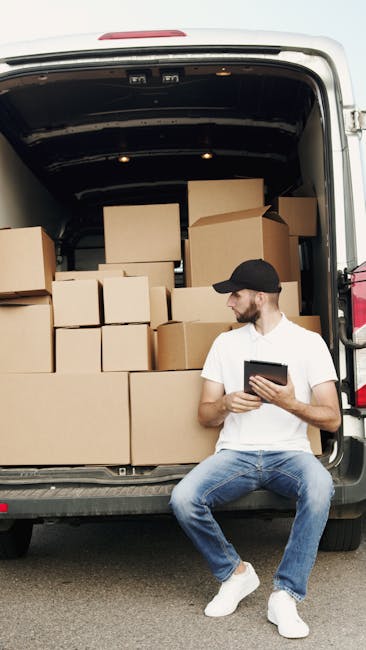 A man in casual clothing, including a white t-shirt, jeans, white sneakers, and a black cap, is sitting on the pavement beside an open van loaded with numerous cardboard boxes of various sizes. The boxes are stacked inside the vehicle, which is positioned on a driveway or street, and some are arranged on the pavement near the man. The interior of the van reveals a dark space with additional boxes, suggesting an ongoing home relocation or furniture transport process. The man is holding a tablet or mobile device, possibly coordinating the packing or loading activities. The scene is set in daylight, with natural lighting illuminating the boxes, the man, and the van, reflecting a professional move managed by Man with Van Ardleigh Green, which offers removals and moving services, including packing, loading, and transportation.