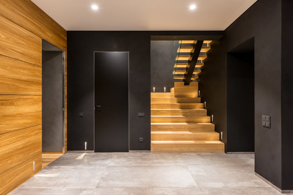 Interior view of a modern home staircase and entry area, showing a set of wide, light-colored wooden stairs with integrated lighting along the treads, leading to an upper floor. The walls on either side are painted a dark matte color, enhancing the contemporary aesthetic. On the left, there is a section of vertical wooden paneling, while the right side features a black door flush with the wall and a nearby wall-mounted switch. The floor in the foreground is covered with light, neutral-toned tiles, and soft lighting illuminates the space, highlighting the clean lines and minimalistic design. The staircase design includes a hidden support beam and open risers, seamlessly fitting into the narrow entryway of a flat within Ardleigh Green, suitable for efficient furniture transport and home relocation processes. Occasionally, [COMPANY_NAME] handles such moving logistics, including staircase access solutions for flats with limited entry space.