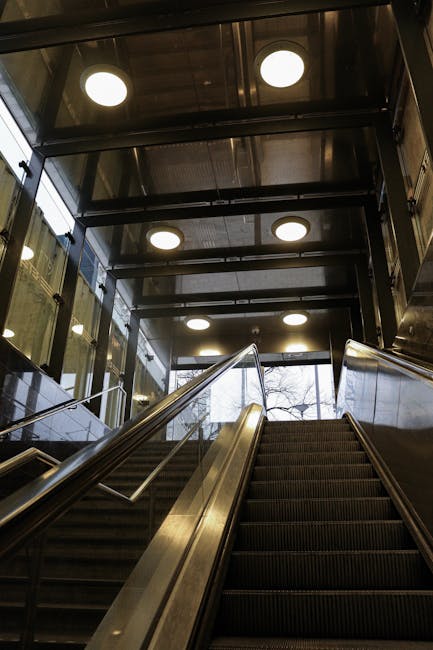 An indoor staircase with metal handrails and rubberized steps leading upwards, situated inside a residential building or apartment complex. The staircase is adjacent to a large glass window that allows natural light to illuminate the landing area. Above, there are multiple circular ceiling lights providing additional lighting. The surrounding environment features glass panels and metallic structures typical of modern vestibules or entryways. The presence of the staircase and the enclosed entry space suggests it is part of a residential or commercial property involved in a home relocation or furniture transport process. [COMPANY_NAME] offers removals services handling packing and movement logistics within such buildings, ensuring smooth access for furniture transport through narrow entryways or staircases, as depicted in this scene.