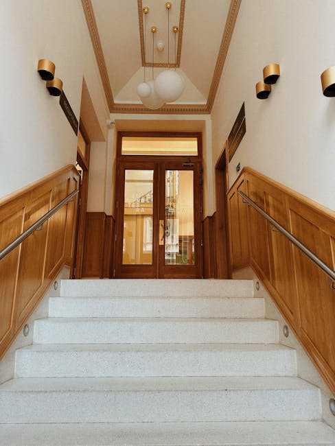 Interior view of a modern home staircase and entry area, showing a set of wide, light-colored wooden stairs with integrated lighting along the treads, leading to an upper floor. The walls on either side are painted a dark matte color, enhancing the contemporary aesthetic. On the left, there is a section of vertical wooden paneling, while the right side features a black door flush with the wall and a nearby wall-mounted switch. The floor in the foreground is covered with light, neutral-toned tiles, and soft lighting illuminates the space, highlighting the clean lines and minimalistic design. The staircase design includes a hidden support beam and open risers, seamlessly fitting into the narrow entryway of a flat within Ardleigh Green, suitable for efficient furniture transport and home relocation processes. Occasionally, [COMPANY_NAME] handles such moving logistics, including staircase access solutions for flats with limited entry space.